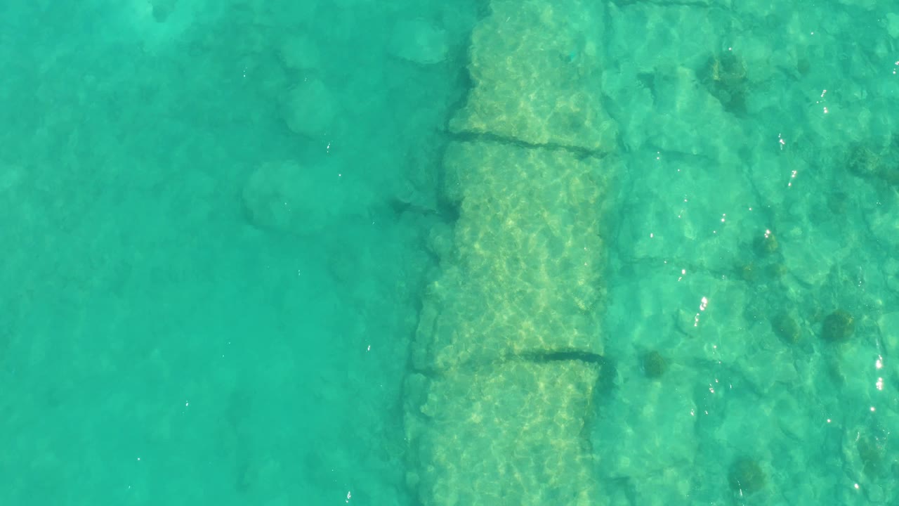 An overhead Cabo Pulmo frame shows green-blue water, a vertical submerged stone form, scattered rocks to the side, faint fish movement, and no visible people.