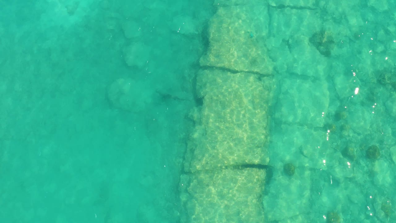 Turquoise water in Cabo Pulmo reveals rectangular stone sections below the surface, with darker rock patches, small fish near the edge, and no visible people.