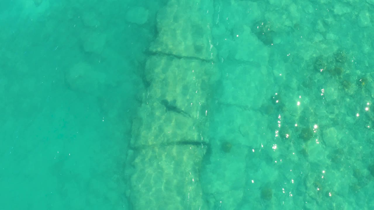 Overhead Cabo Pulmo view with a submerged stone structure left of center, bright reflected points on the water, and no visible people.