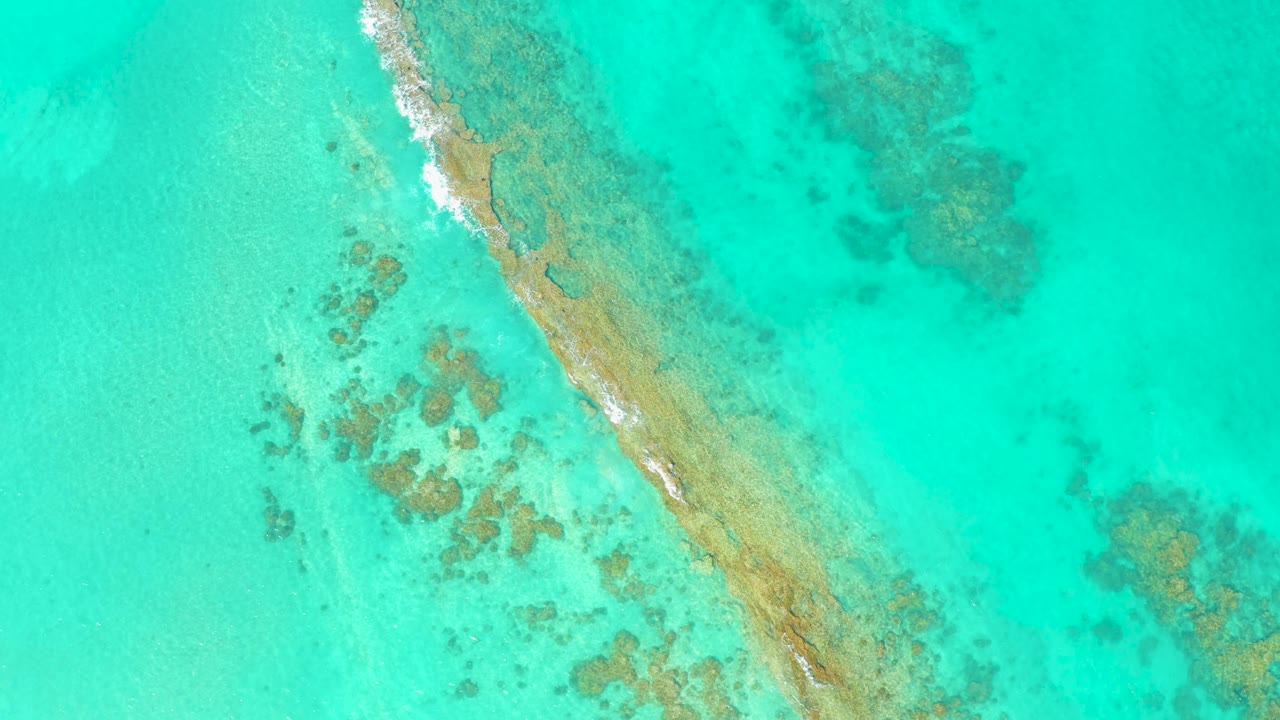 Cabo Pulmo overhead view with the sandbar still running diagonally as faint foam gathers along one edge and darker coral or rock patches sit in the shallow water, with no visible people.
