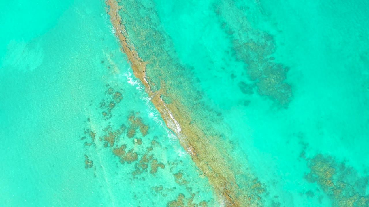 Cabo Pulmo aerial frame with a narrow diagonal reef or sandbar line, small white waves breaking on its edge, pale turquoise water, and no visible people.
