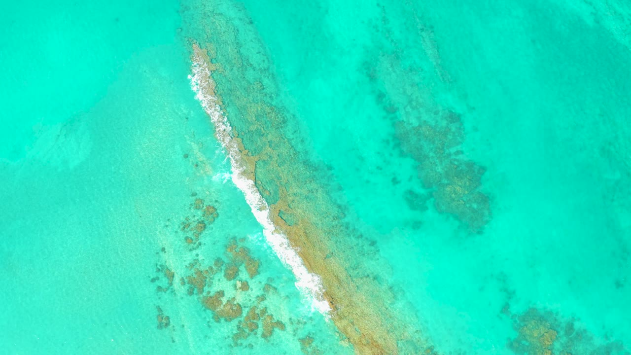 Overhead view in Cabo Pulmo, Mexico showing clear turquoise water, a diagonal tan sandbar or reef line, small white foam on the left edge, and no visible people.