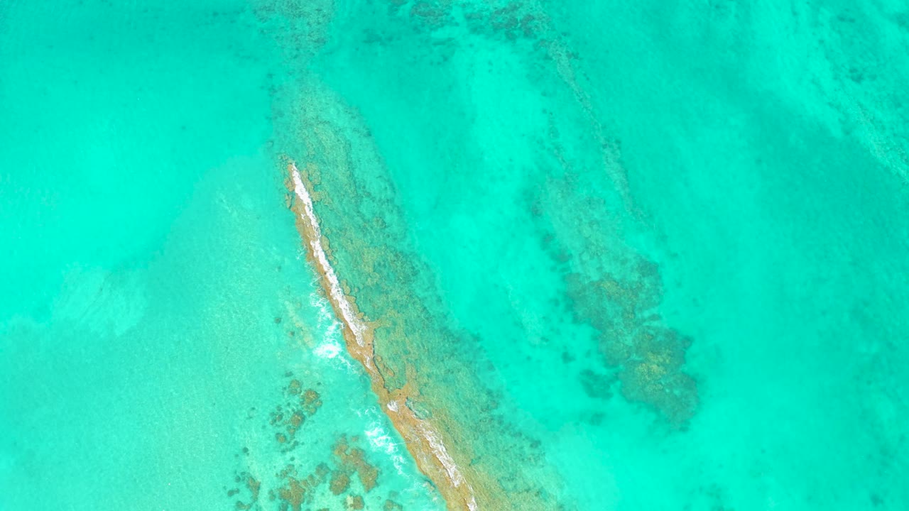 High overhead view in Cabo Pulmo with a narrow sandbar lower in frame, shallow aqua water, darker coral patches, and no visible people.