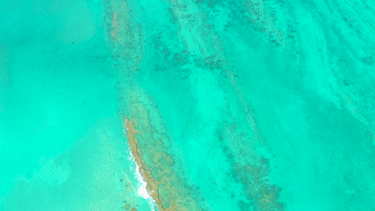 Aerial Cabo Pulmo frame with the sandbar slightly lower in view, darker submerged seabed streaks to the right, pale shallow water to the left, and no visible people.