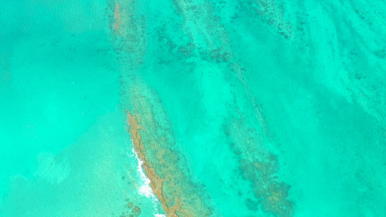 Cabo Pulmo aerial frame showing the sandbar edge more centered, with a small white wave break along the left side and soft underwater bands visible through the turquoise water, no visible people.