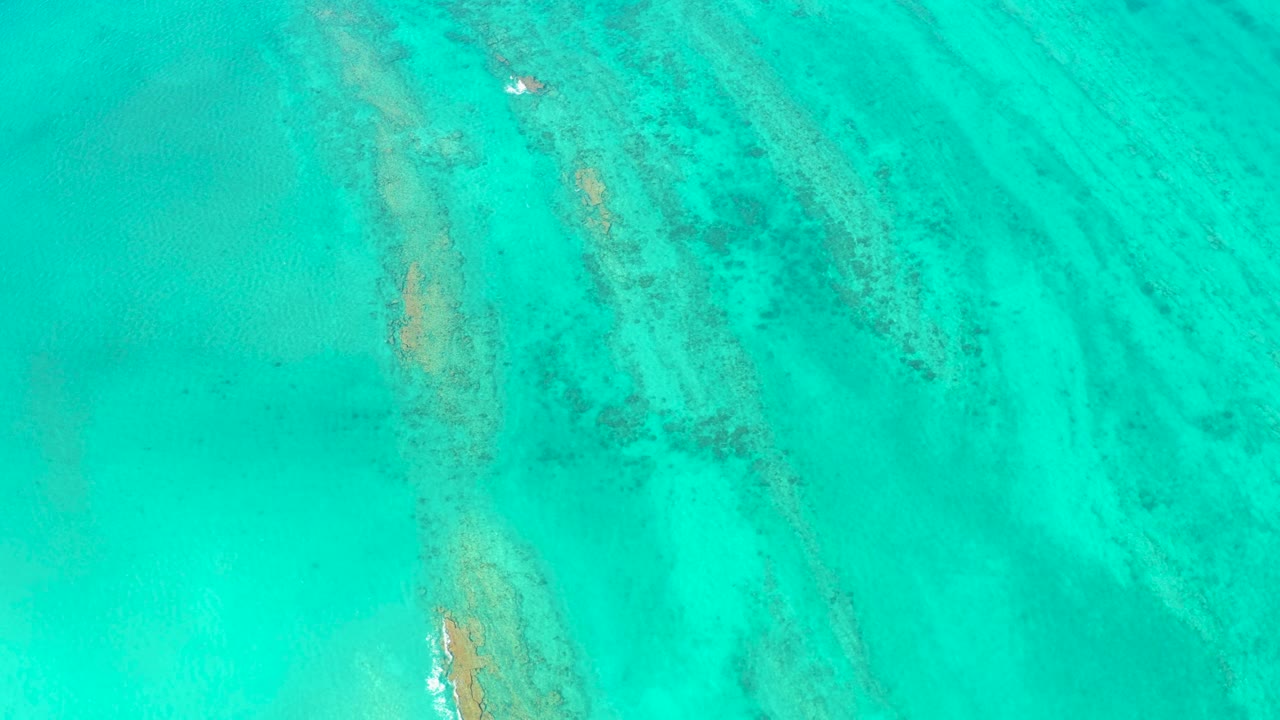 Aerial frame over Cabo Pulmo turquoise sea, showing dark reef-like streaks across the right side and a pale surface speck near the upper left, no visible people.