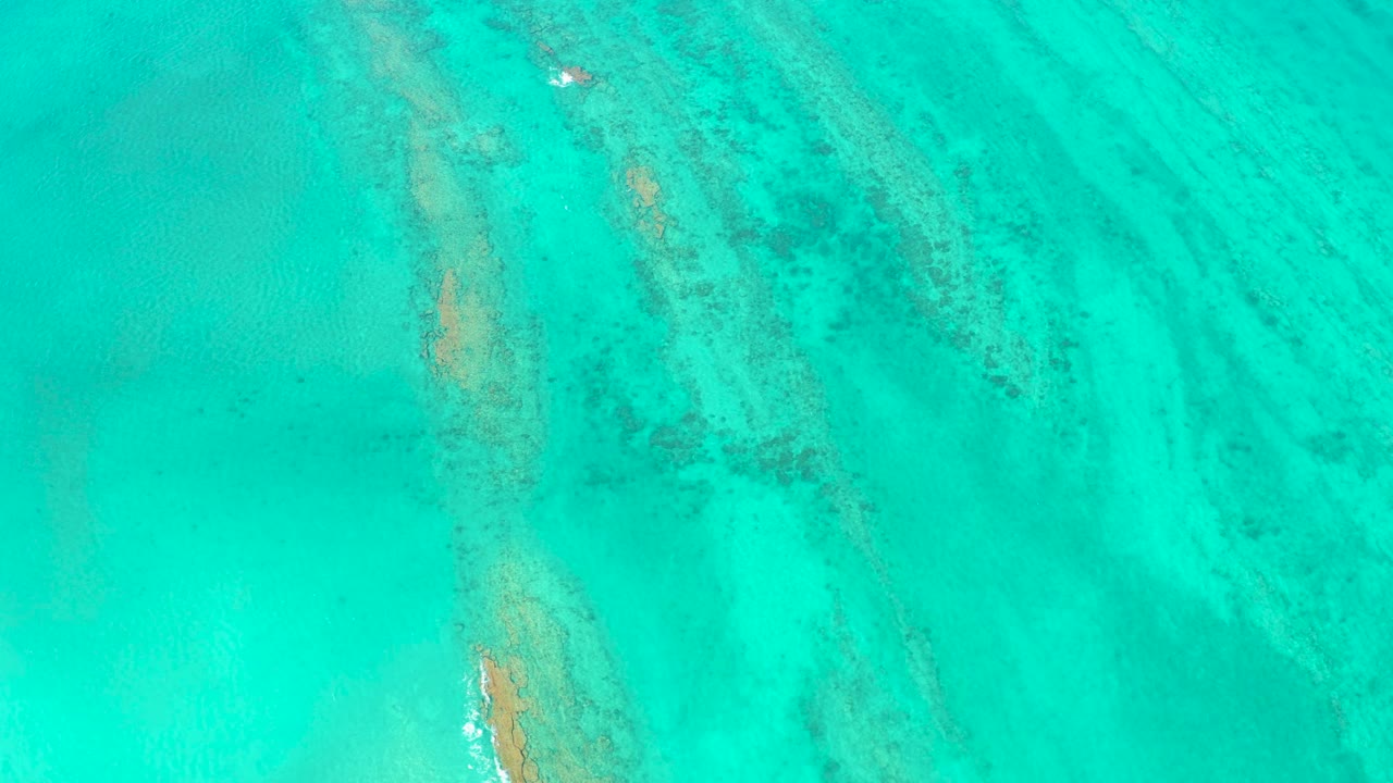 Mid-clip overhead view in Cabo Pulmo with turquoise water, a vertical tan submerged formation near the center-left, darker patches to the right, and no visible people.