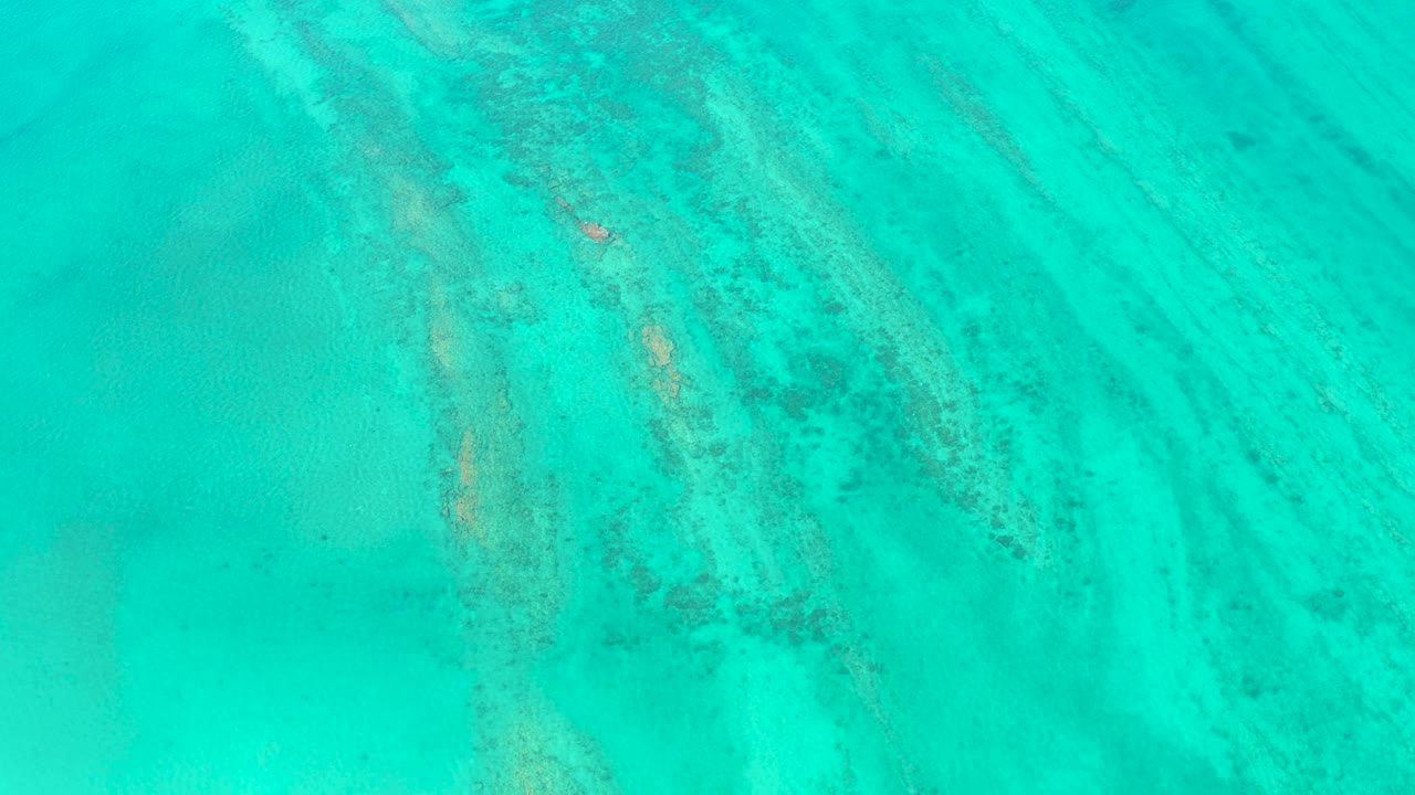 Overhead view in Cabo Pulmo, Baja California Sur of turquoise ocean water with faint underwater sand and reef streaks, a small white boat near the patterned water, and no visible people.