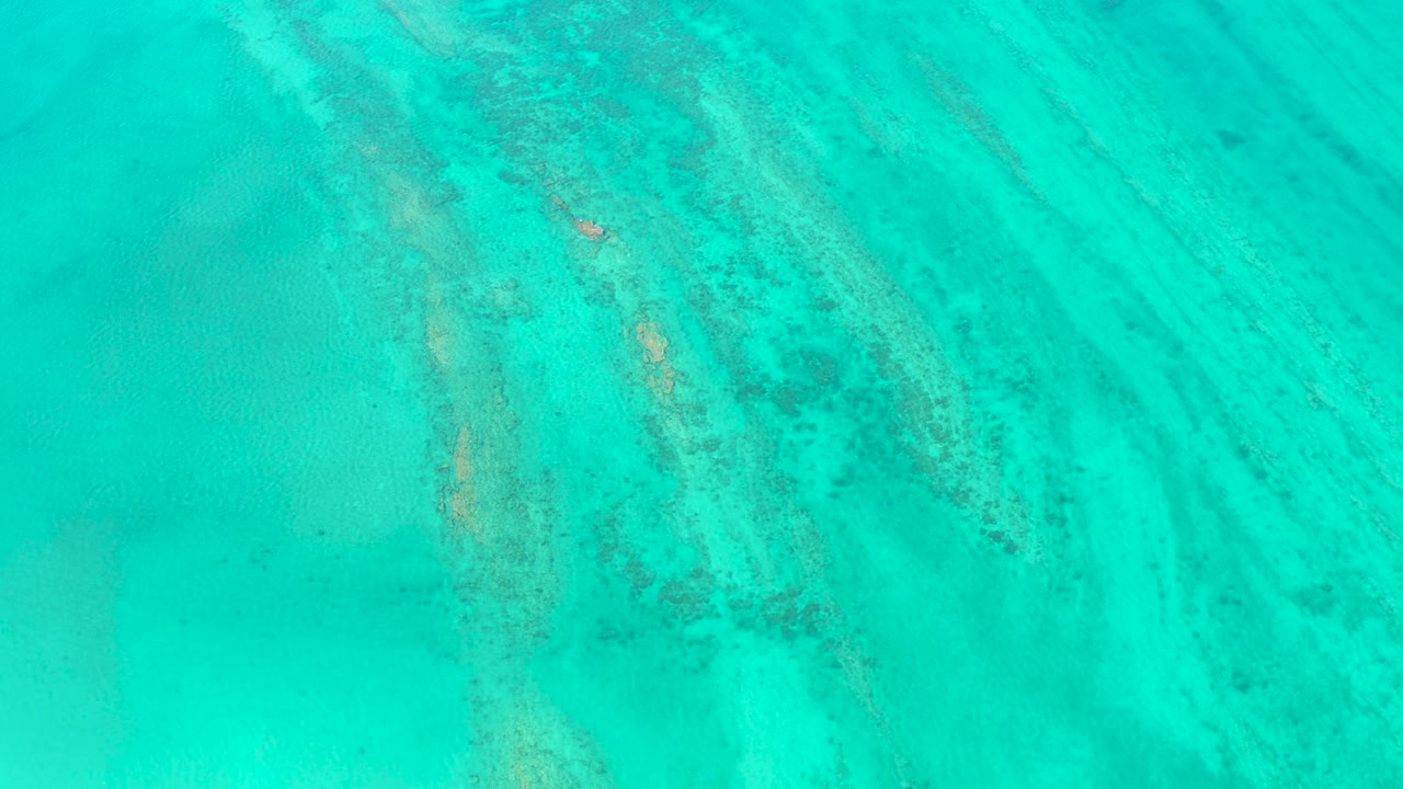 Aerial frame from Cabo Pulmo showing clear blue-green ocean, diagonal darker underwater marks, a small white boat slightly farther along the water path, and no visible people.