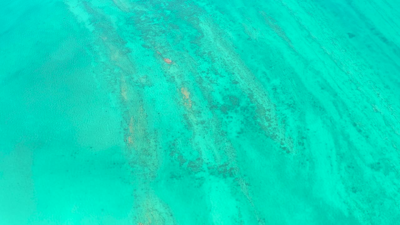Top-down Cabo Pulmo ocean view with pale sand bands, scattered darker underwater patches, the small white boat set against turquoise water, and no visible people.