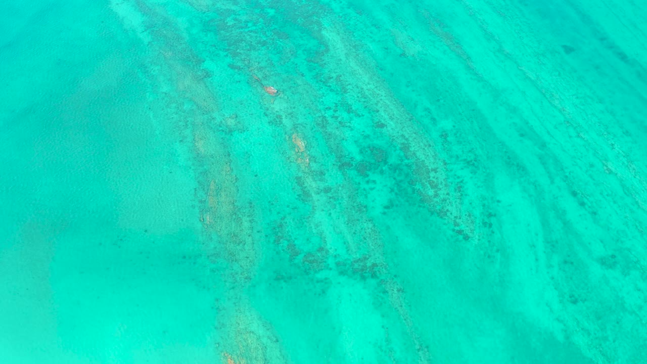 Drone-style overhead frame in Cabo Pulmo with reef-like markings under bright turquoise water, a small white boat crossing the textured area, and no visible people.