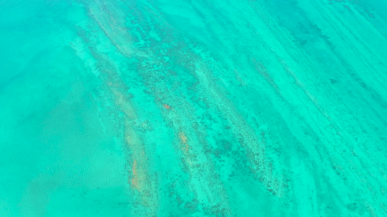 Blue-green shallow water in Cabo Pulmo covers a visible ocean floor with diagonal sand lines, mottled reef-like patches, and no visible people.