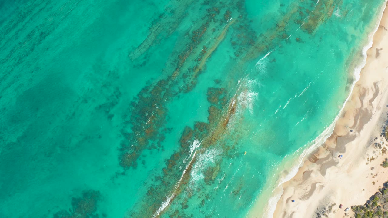 Overhead view of turquoise water meeting a pale sandy beach in Cabo Pulmo, with darker reef patches below the surface and no visible people.