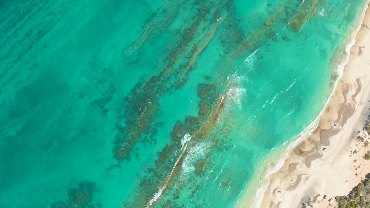 Aerial frame over Cabo Pulmo shoreline, where clear teal water covers dark seabed patches and small white foam streaks approach the sand with no visible people.
