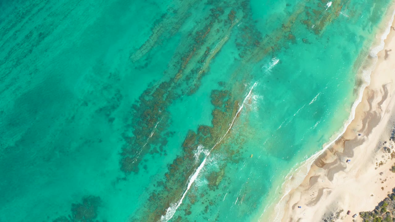 A white foam band advances through turquoise shallows toward the sandy Cabo Pulmo shore, with darker underwater patches on the left and no visible people.