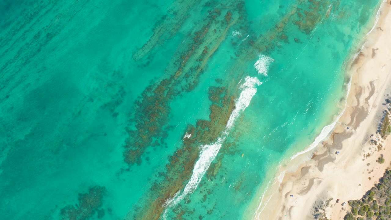 Overhead view in Cabo Pulmo with turquoise water covering dark reef patches, a pale sandy beach at right, small white surf lines, and no visible people.