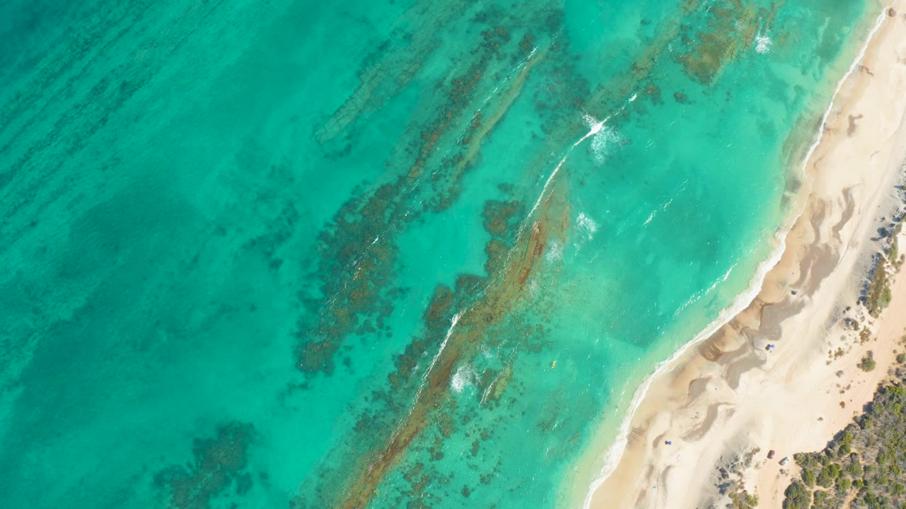 Aerial frame of Cabo Pulmo shoreline with the white foam streak slightly farther along the shallow break, dark underwater patches across the water, pale beach at right, and no visible people.