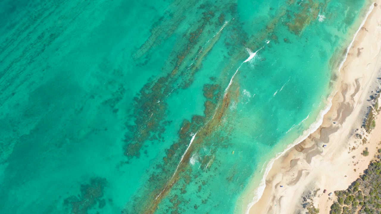 Top-down Cabo Pulmo view with bright white foam near the upper right shallows, a long darker underwater band through the center, pale sand on the right, and no visible people.
