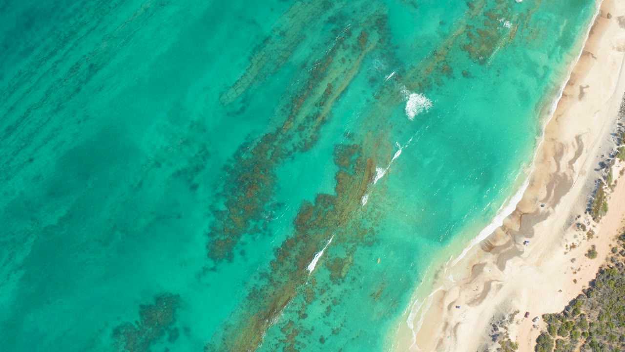 Cabo Pulmo shoreline from above, with broad turquoise water, darker reef patches through the center, pale sand at right, a small breaking white wake, and no visible people.