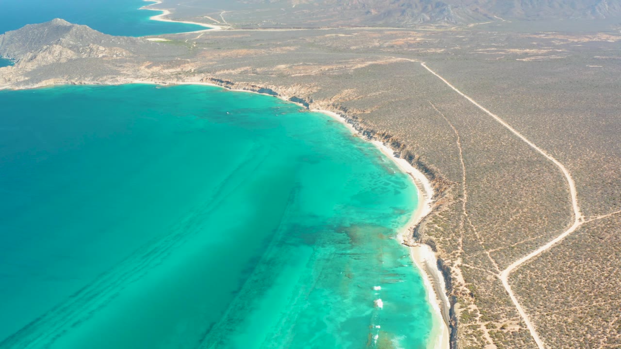 High drone frame over Cabo Pulmo showing the turquoise bay closer in the foreground, a narrow rocky beach, branching pale roads across scrub desert, distant mountains, and no visible people.