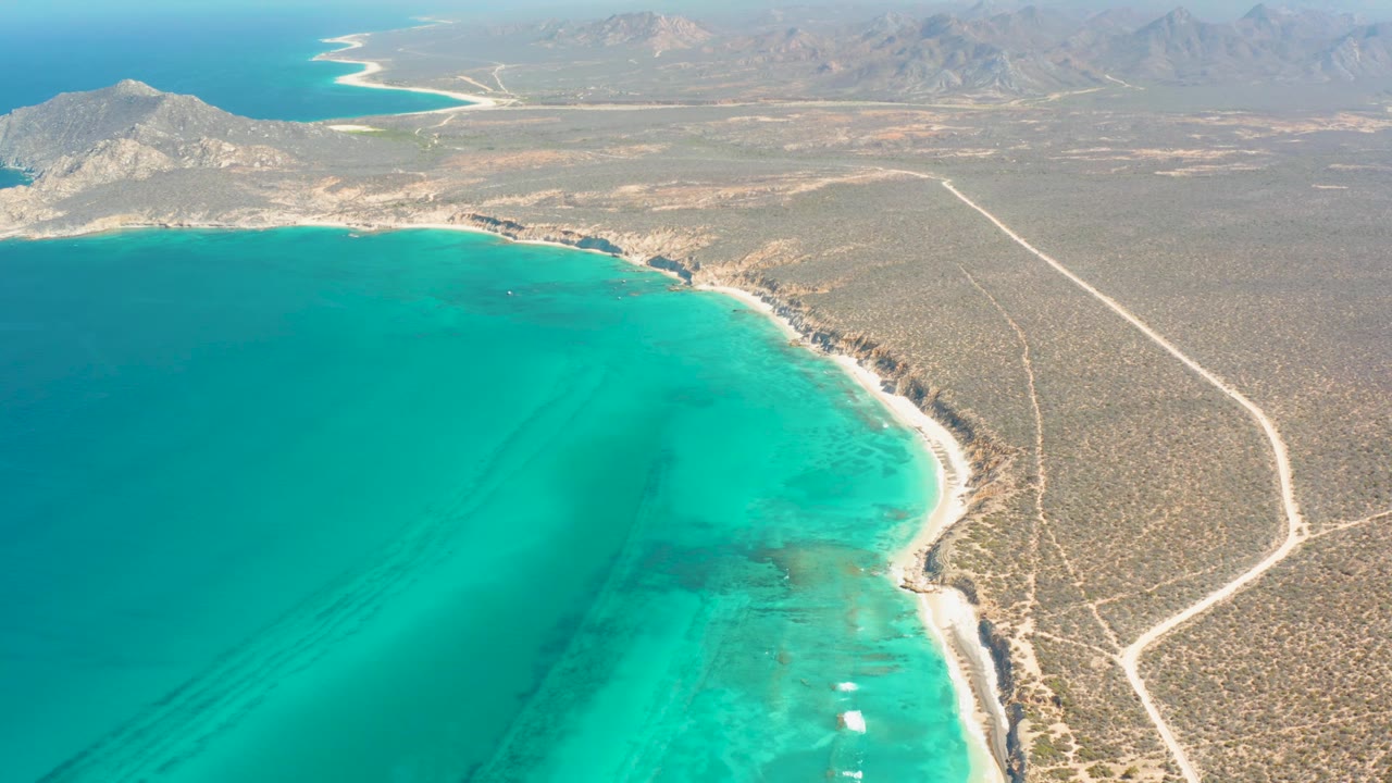 Mid-clip aerial over Cabo Pulmo with open turquoise water at left, a curving beach edge, scrub-covered desert at right, a pale road bending inland, distant mountains, and no visible people.