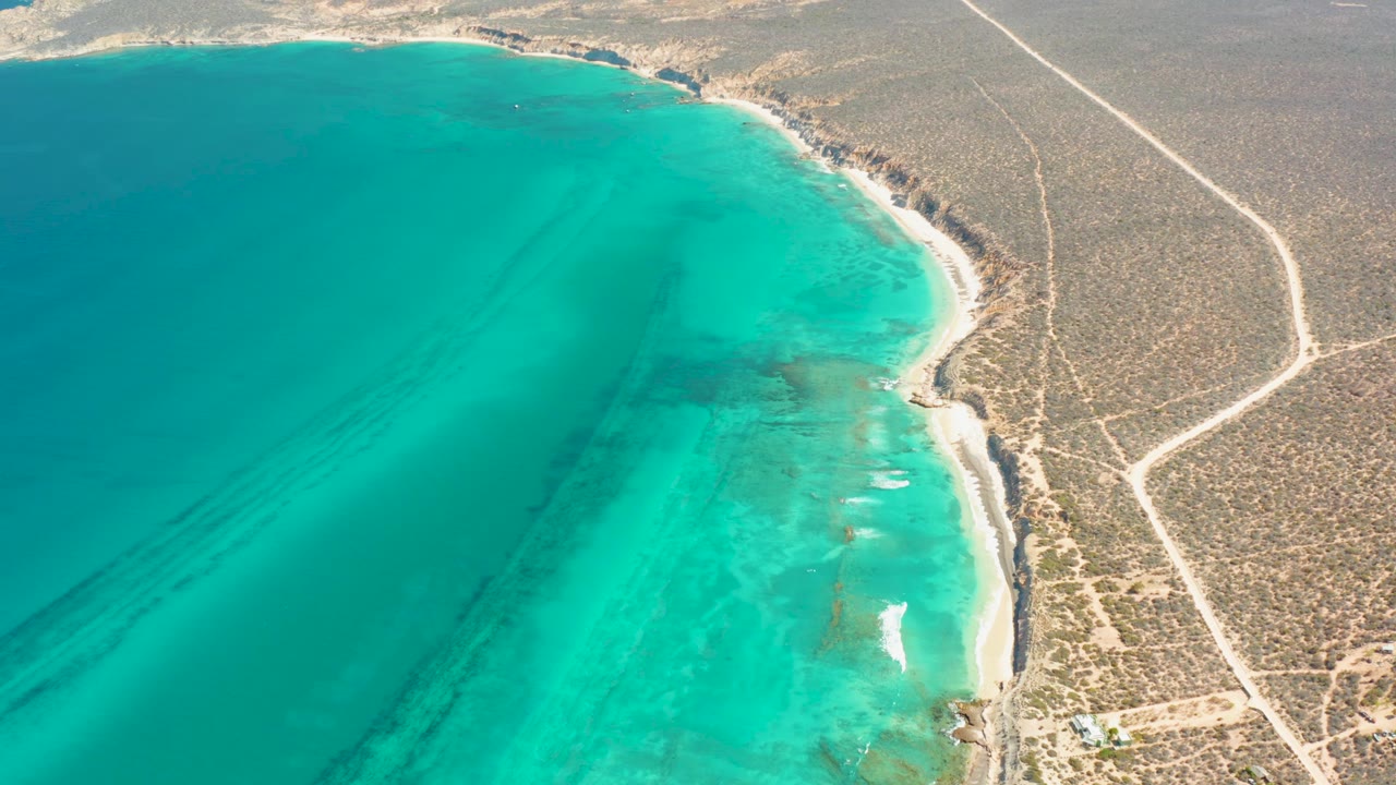 Aerial view of Cabo Pulmo coastline with broad turquoise water, darker shallow patches near shore, a narrow beach, inland desert roads, and no visible people.