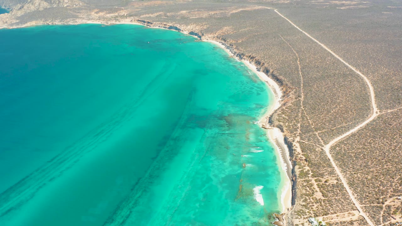 Cabo Pulmo aerial frame with turquoise ocean dominating the left, a bright sandy strip along the right edge of the water, dry desert beyond it, and no visible people.
