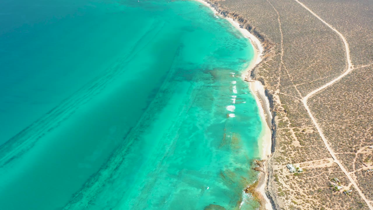 Aerial frame over Cabo Pulmo with turquoise water on the left, a pale shoreline on the right edge of the bay, dry scrubland and dirt roads inland, and no visible people.
