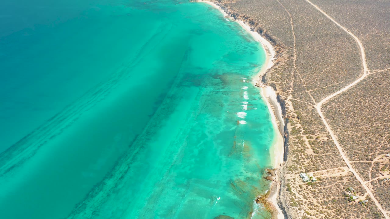 Aerial frame in Cabo Pulmo with a turquoise bay, small white surf marks near the shore, brown arid terrain, pale unpaved roads, small inland structures, and no visible people.