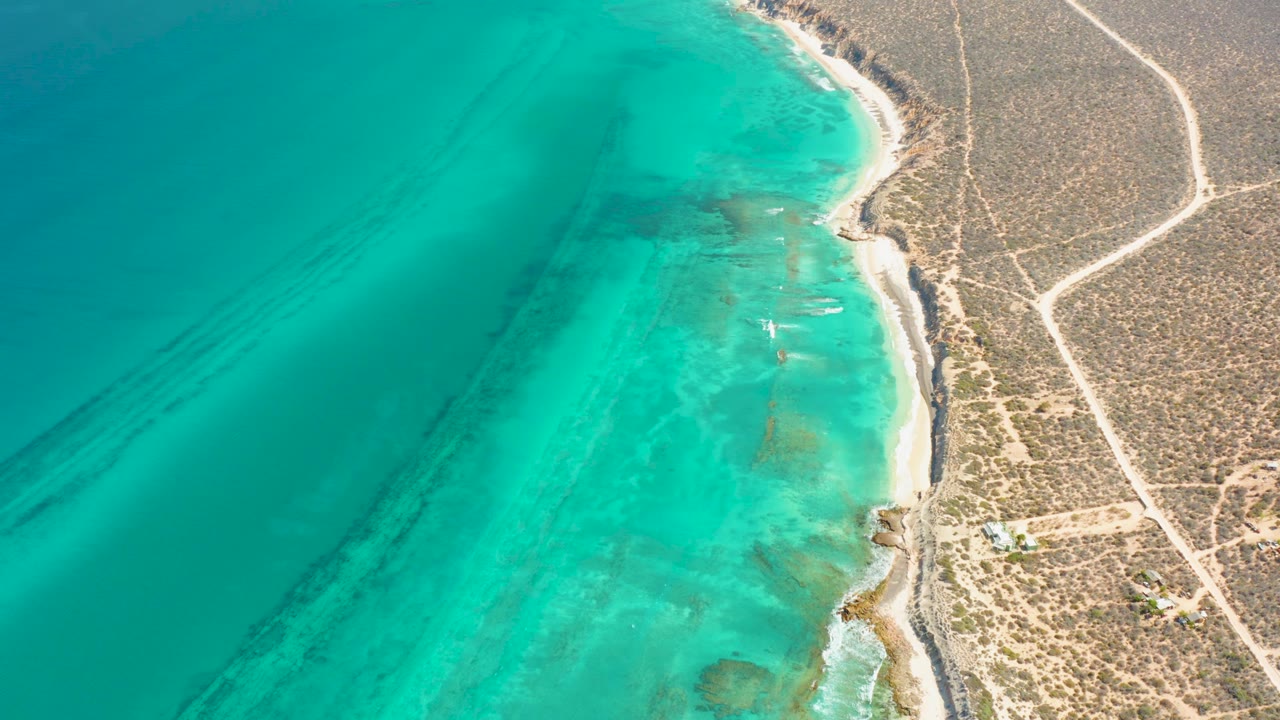 Second aerial frame over Cabo Pulmo with shallow turquoise water, darker underwater bands, a narrow beach, and pale dirt roads across dry land, with no visible people.