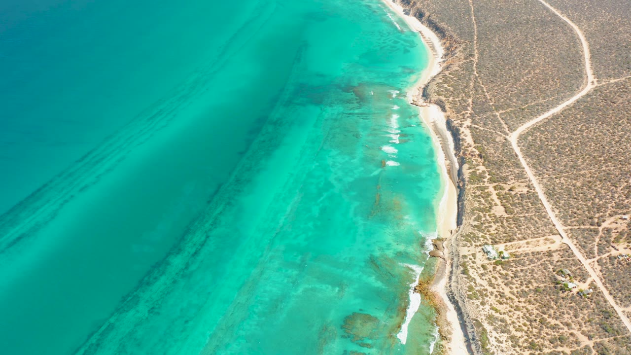 Aerial view in Cabo Pulmo with shallow turquoise water, small white surf lines, dry scrubland, pale roads, and no visible people.