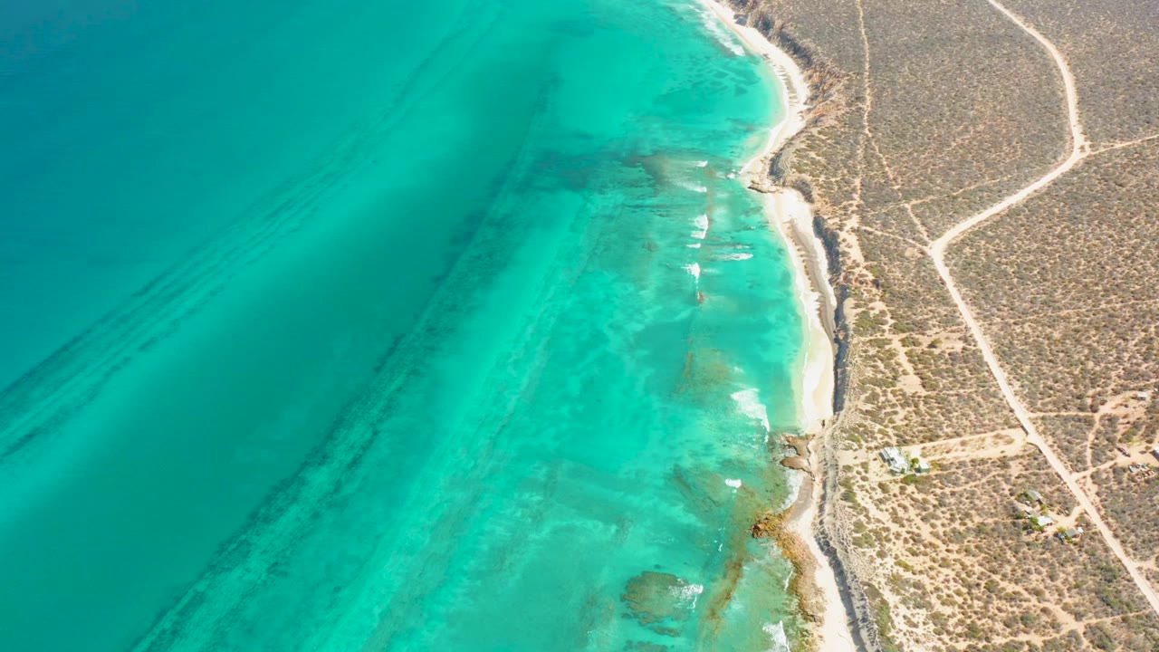 Aerial frame over Cabo Pulmo showing broad turquoise shallows, small white wave marks along the beach, dry brush inland, pale dirt tracks, and no visible people.