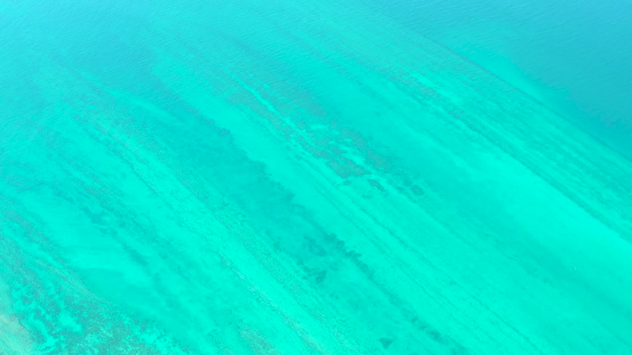 Bright turquoise water in Cabo Pulmo covers pale sand with soft sunlight ripples and a darker patch near the lower area, with no visible people.