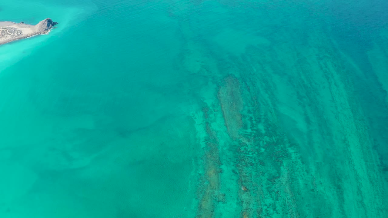 A Cabo Pulmo aerial frame shows the island slightly farther inside the upper left, with teal water and darker seabed texture running down the right side, no visible people.