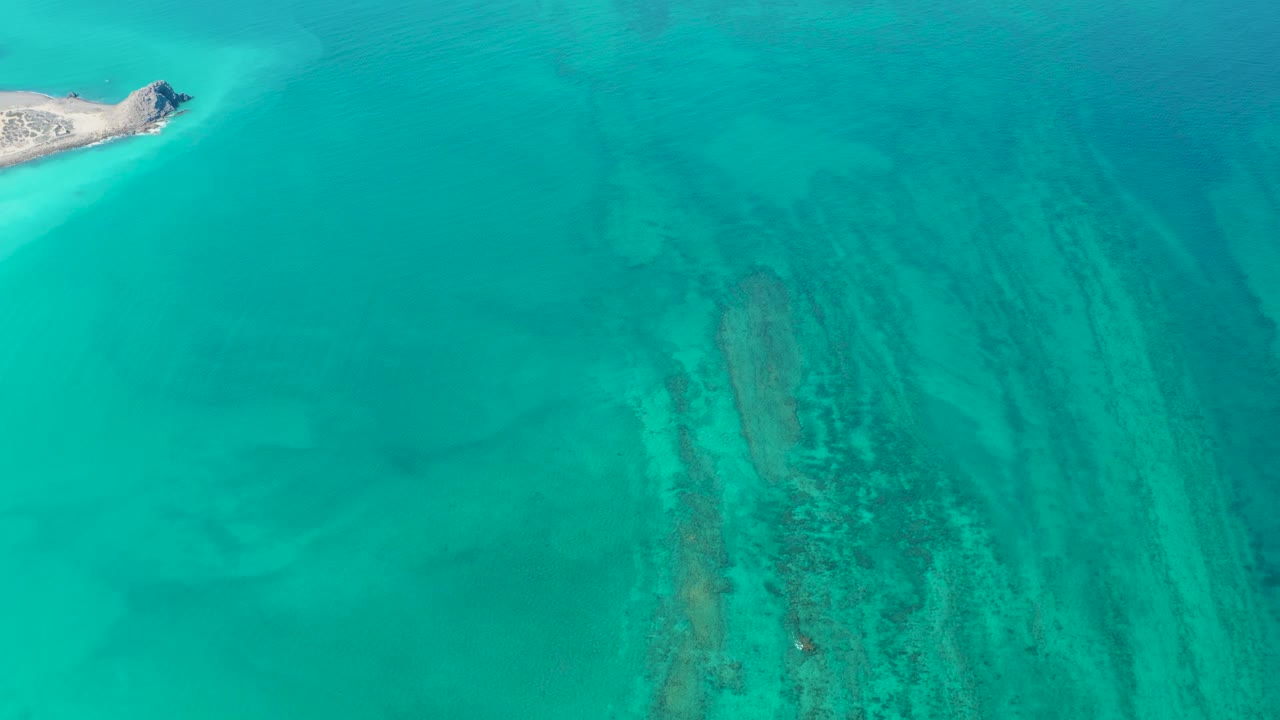 The island sits near the upper left of the Cabo Pulmo frame while clear turquoise water reveals darker reef streaks and pale sand across the right half, with no visible people.