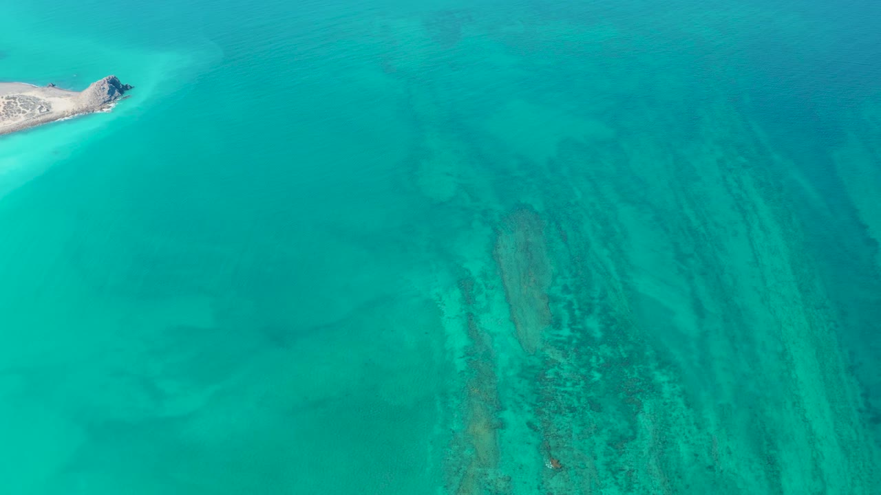 Mid-clip overhead view in Cabo Pulmo shows the island near the left edge, open teal water across the frame, and submerged coral and sand patterns, no visible people.