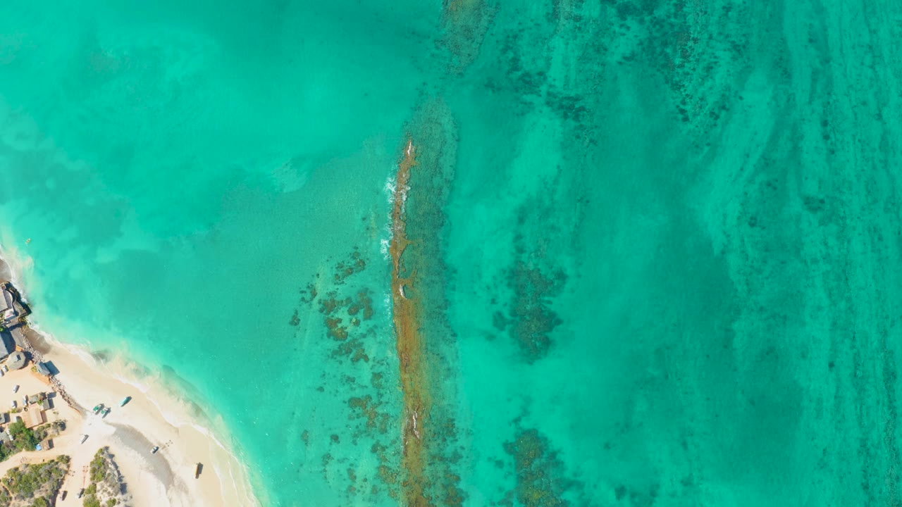 Aerial view over Cabo Pulmo water where the small boat remains near the lower left beach edge and a thin reef line runs vertically through the frame, with no visible people.