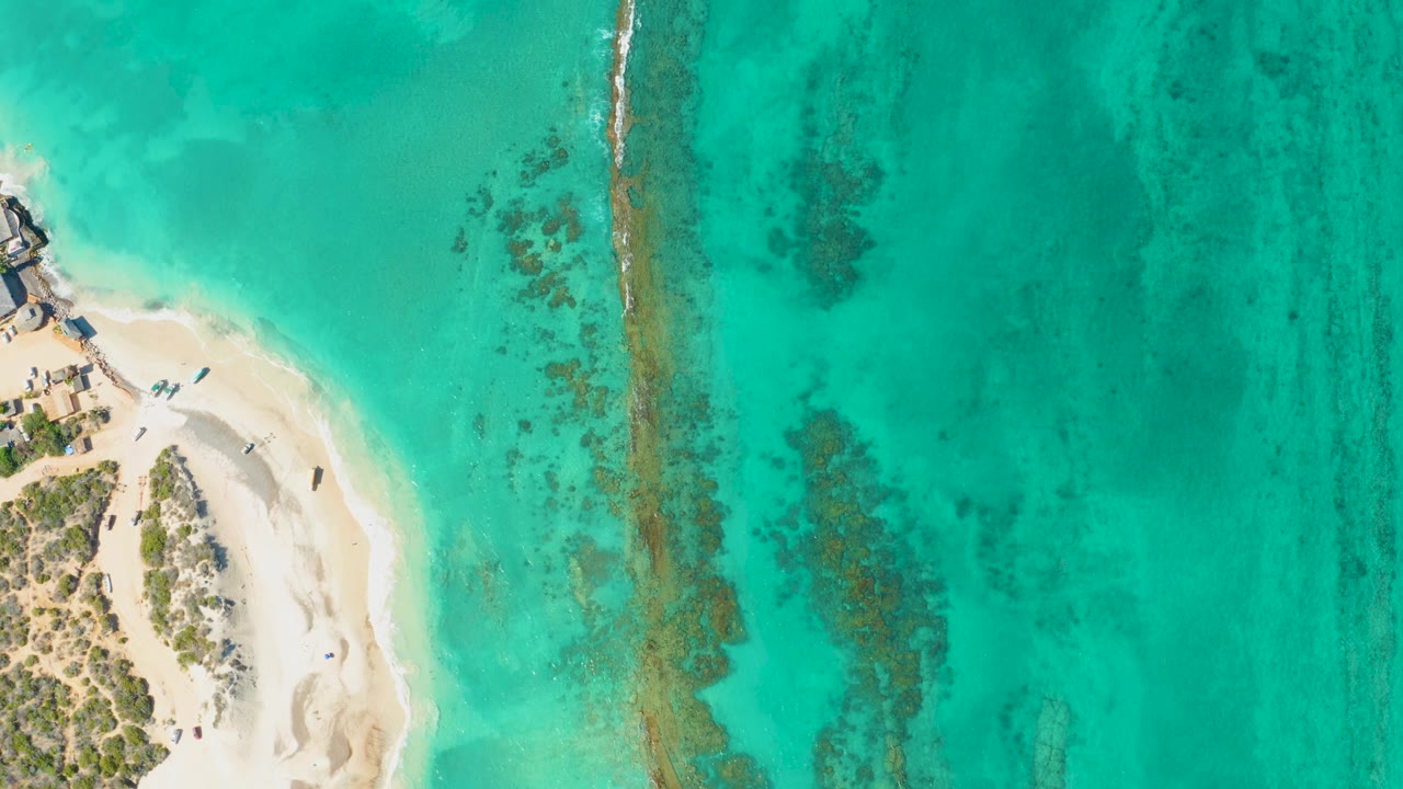 Aerial shoreline with clear blue-green water and pale sand at the left.