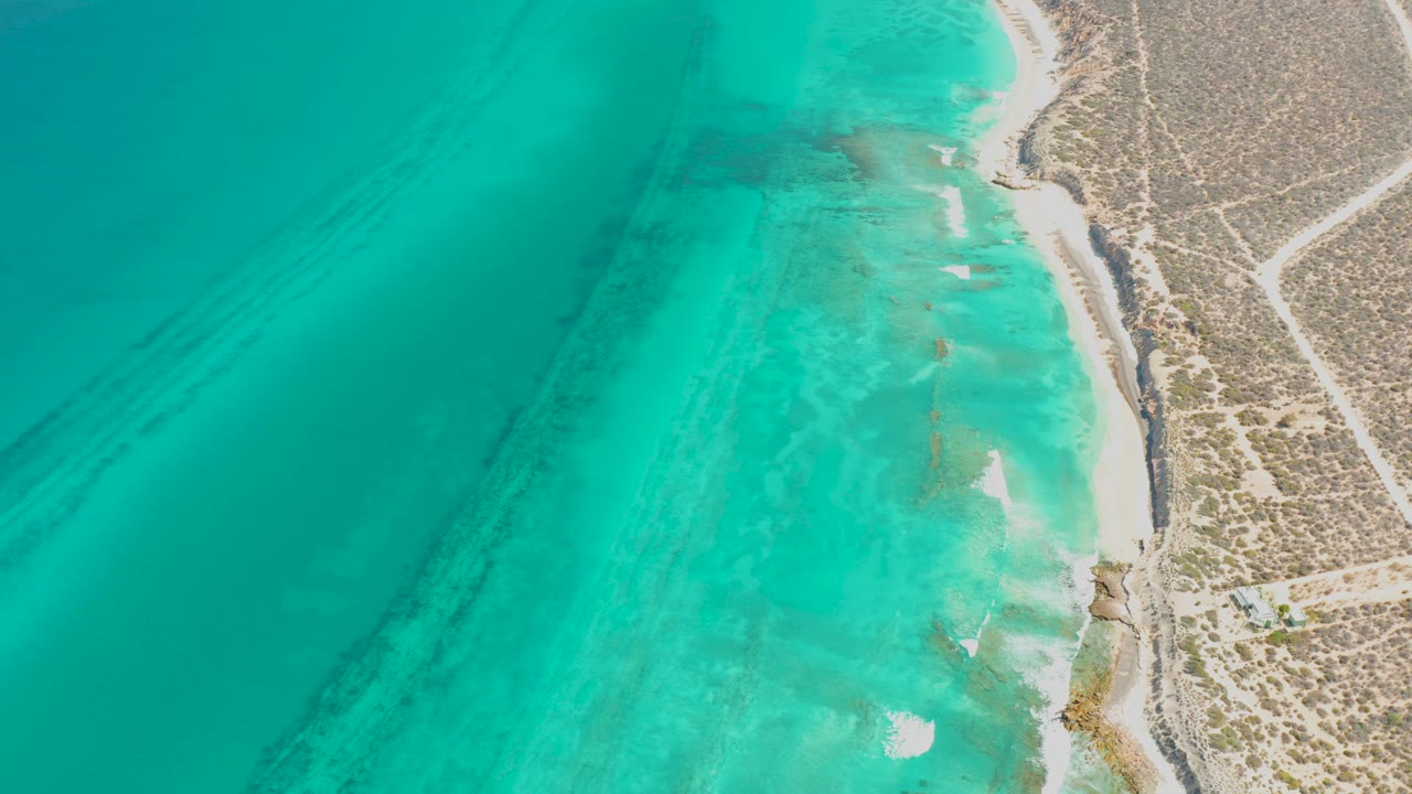Aerial frame of shallow turquoise surf along Cabo Pulmo, with pale sand exposed near the waterline and no visible people.