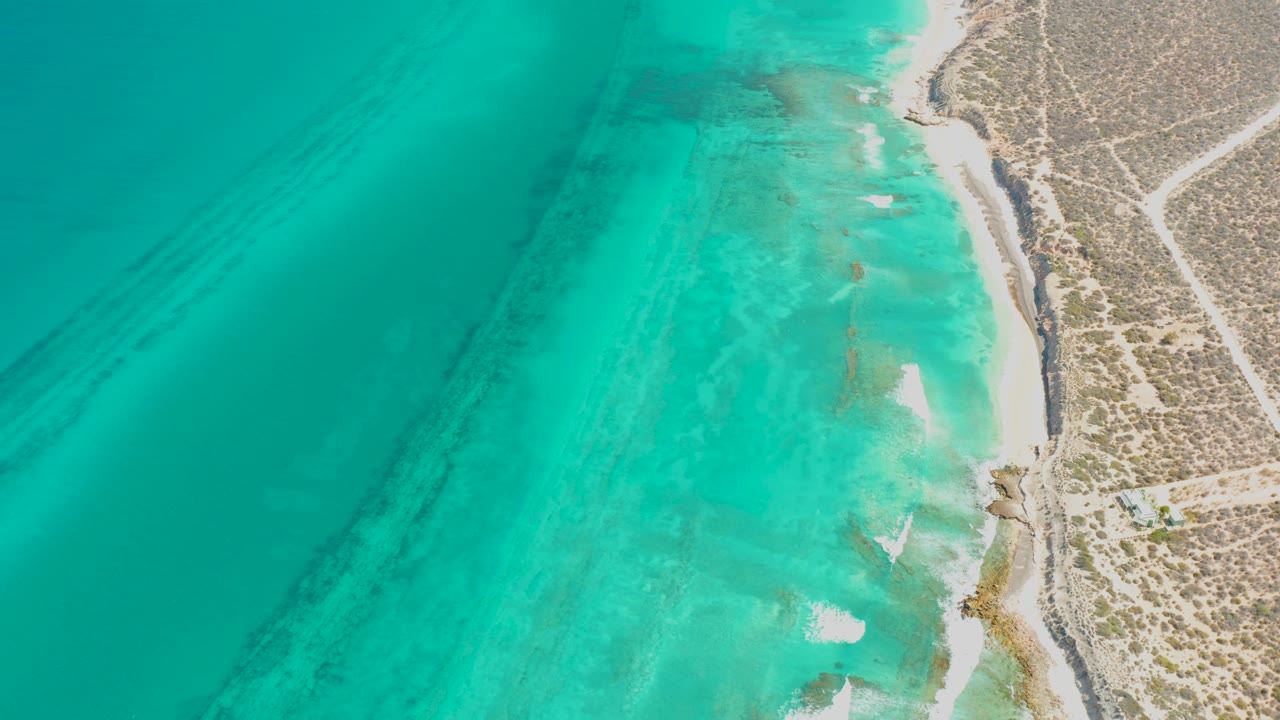 Drone view over clear turquoise water at Cabo Pulmo, where small white waves touch the sandy shore and no visible people appear.