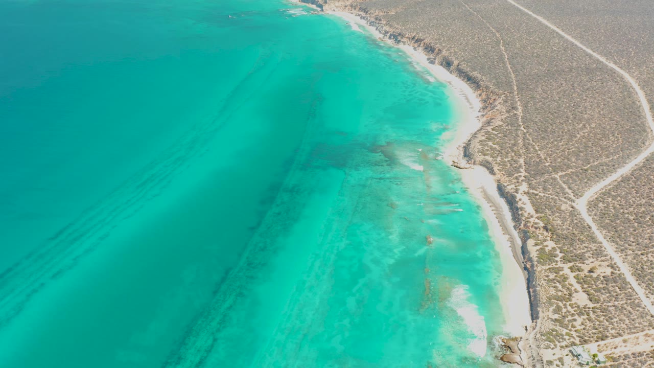 Aerial view over Cabo Pulmo with the shoreline slightly farther along the coast, showing blue-green shallows, pale sand, scrubland, and no visible people.