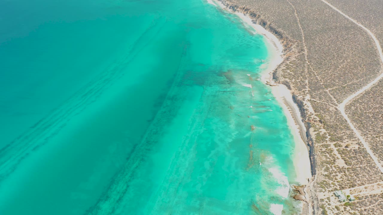 Mid-clip aerial over Cabo Pulmo showing deeper turquoise water at left, pale shallows near the beach, desert scrub inland, and no visible people.
