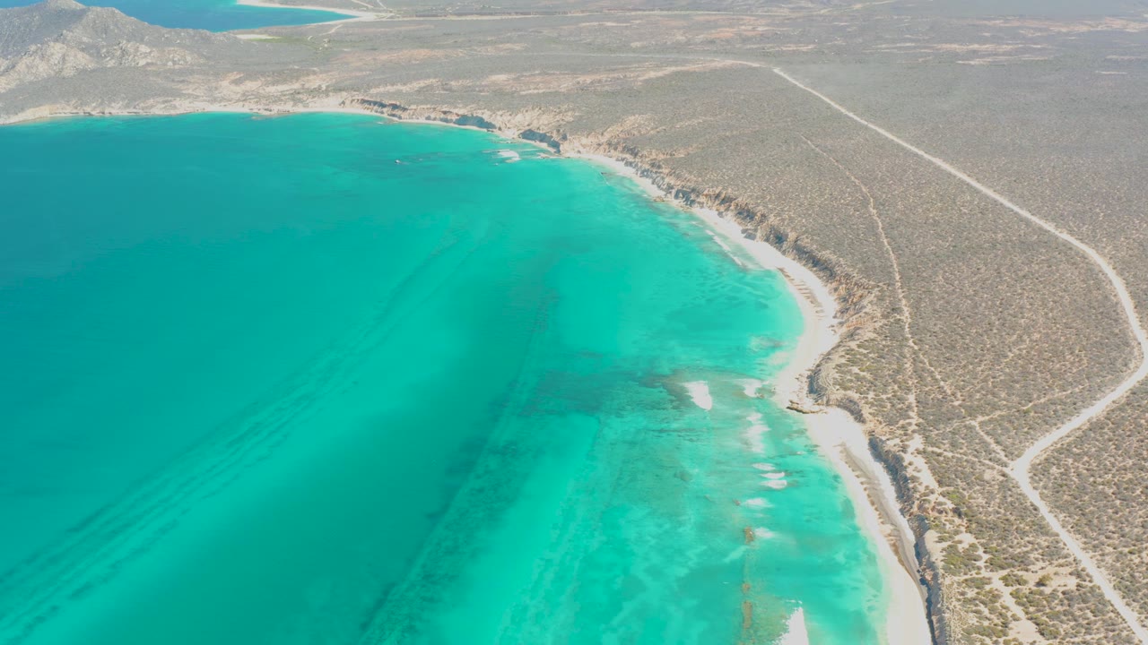 Aerial frame at 0:01 over Cabo Pulmo showing the curved turquoise bay, pale shoreline, scrub-covered desert, a light dirt road, and no visible people.