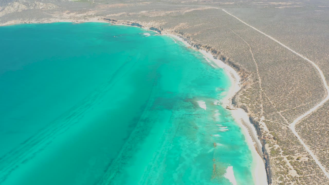 Aerial frame at 0:03 over Cabo Pulmo with blue-green water dominating the left, a narrow pale shoreline below the desert edge, a dirt road on the right, and no visible people.