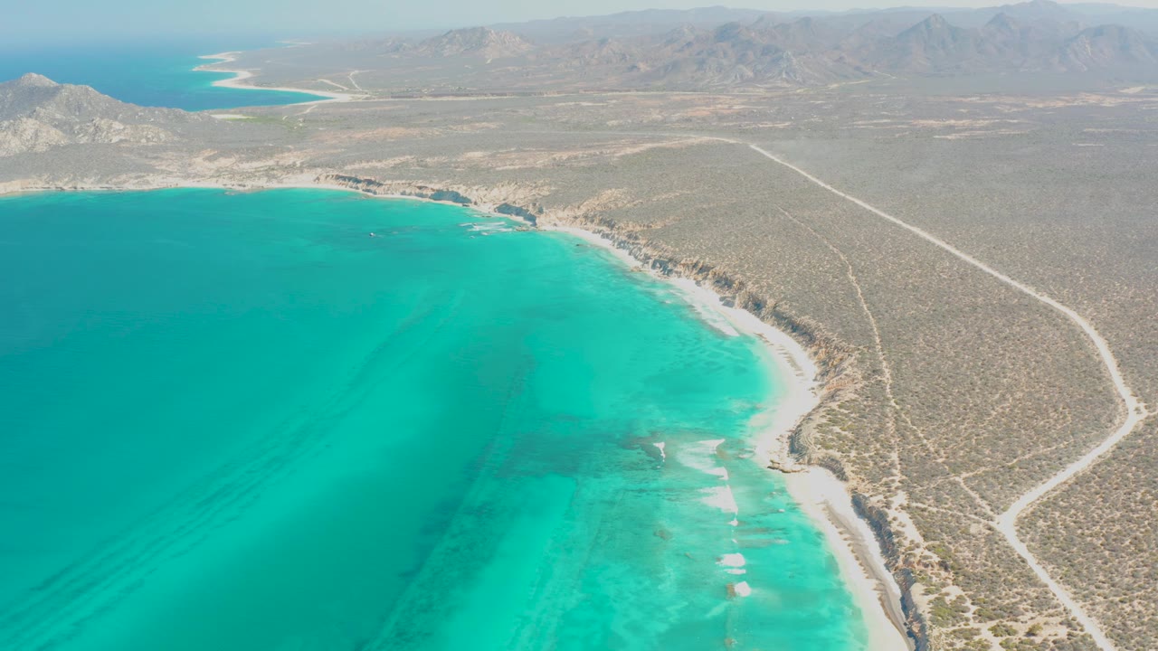 Aerial frame over Cabo Pulmo showing turquoise water on the left, a pale sandy shoreline, desert scrubland, distant hills, and no visible people.