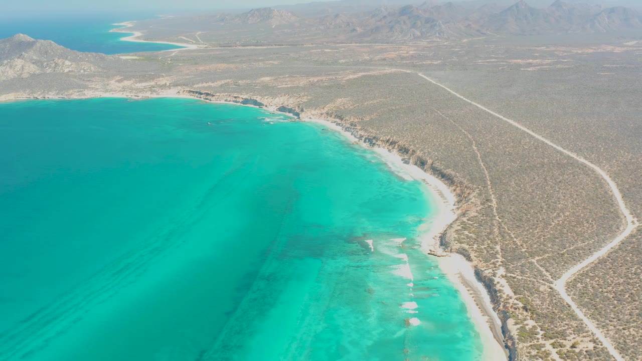High aerial frame of Cabo Pulmo coastline with broad turquoise water, a thin beach line, scrub-covered desert, a pale road crossing inland, distant mountains, and no visible people.