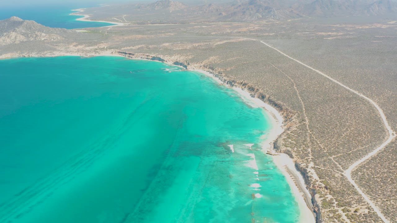 Aerial view over Cabo Pulmo with deeper turquoise water in the foreground, small white breaks near the beach, dry scrubland to the right, a pale road, and no visible people.