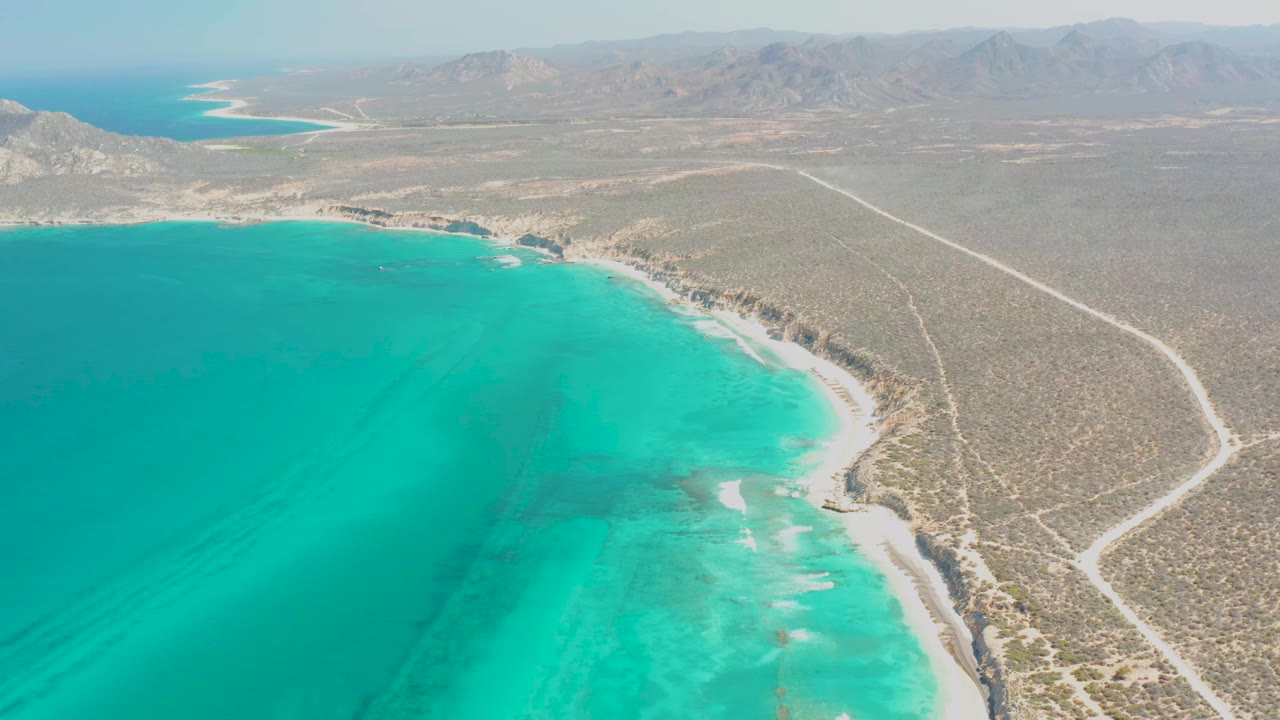 Aerial view of Cabo Pulmo coastline with turquoise ocean at left, a pale sandy shore, dry scrubland, winding dirt roads, distant mountains, and no visible people.