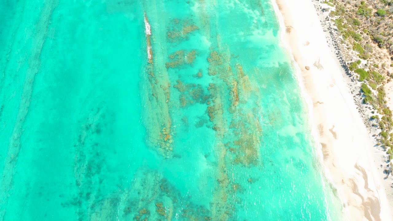 Aerial frame in Cabo Pulmo with the small boat slightly farther along the shallow water, reef and rock shapes visible beneath the surface, white beach at right, and no visible people.