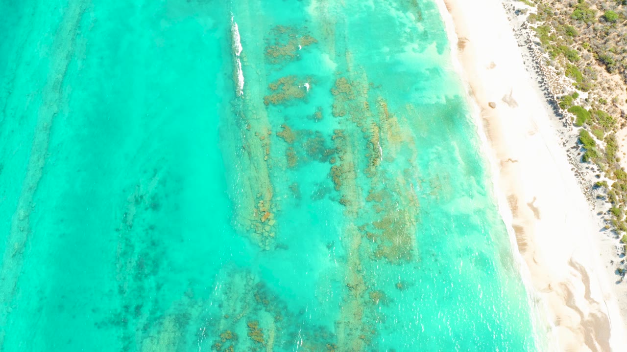 Overhead Cabo Pulmo beach frame with the boat near the upper water, reef patches crossing the shallows, pale sand and scrub vegetation on the right, and no visible people.